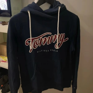 Mörkblå Tommy Hilfiger hoodie S - Snygg mörkblå hoodie från Tommy Hilfiger Denim med stor vit och röd logga framtill. Tröjan har huva med vita snören och känguruficka. Tillverkad i mjuk bomull och polyester, perfekt för chill dagar. Klassisk streetstil med lång ärm.