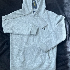 Grå hoodie från Polo Ralph Lauren - Snygg ljusgrå hoodie från Polo Ralph Lauren med klassisk broderad logga på bröstet. Tröjan har huva med snörning, känguruficka och ribbade muddar. Perfekt för en chill och stilren look.