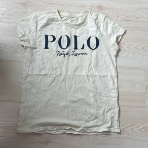 Vit t-shirt från Polo Ralph Lauren - Vit t-shirt från Polo Ralph Lauren med stor blå logga framtill. Klassisk rund halsringning och korta ärmar. Tillverkad i mjuk bomull som känns skön mot huden. Perfekt för en clean och avslappnad stil.