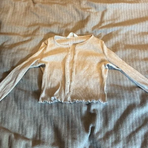 Beige ribbad kofta från H&M - Superfin beige ribbad kofta från H&M med långa ärmar och knappar framtill. Croppad modell med volangkant vid ärmslut och nederkant. Perfekt att slänga över en topp för en chill look.