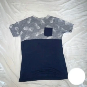 Blå t-shirt med ananasmönster - Snygg t-shirt med mörkblå nederdel och grå överdel med vita ananasmönster. Bröstficka i mörkblått som matchar nederdelen. Perfekt för dig som gillar en chill och lekfull stil.