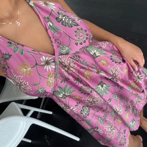 Rosa blommig halterneckklänning - Supersöt rosa kortklänning med blommigt mönster i grönt, vitt och guld. Klänningen har öppen rygg och knyt i nacken samt midjan. Perfekt flowy passform och v-ringad framtill. Materialet känns lätt och svalt, perfekt för varma dagar.