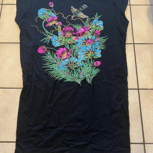 Svart klänning med färgglada blommor - Svart långklänning med stort färgglatt blommönster i blått, rosa och grönt samt en fågel upptill. Klänningen har ärmlös design och raka linjer, perfekt för dig som gillar unika prints och vill sticka ut. 100% bomull 