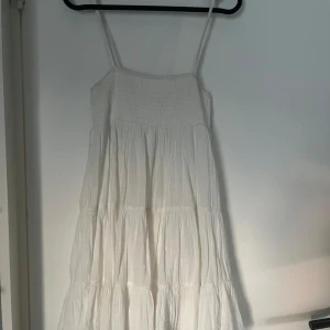 Vit sommarklänning från Zara  - Supersöt vit klänning från Zara i storlek XS. Klänningen har smock upptill, tunna axelband och volangig kjol i flera lager. Perfekt för varma dagar och har en luftig känsla tack vare bomullsmaterialet.