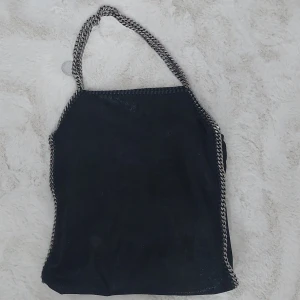 Svart axelväska från Stella McCartney - Snygg svart axelväska från Stella McCartney med kedjedetaljer längs kanterna och axelremmen. Väskan har en mjuk, slouchy form och är tillverkad i syntetmaterial. Perfekt för dig som gillar en cool och trendig look.