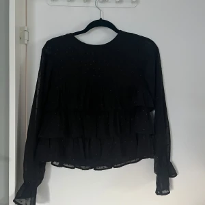 Svart volangblus från Gina Tricot - Supersnygg svart blus från Gina Tricot med volanger och diskreta prickar. Långärmad modell i tunn, luftig polyester med transparenta ärmar och volangdetaljer framtill. Perfekt för dig som gillar trendiga och feminina plagg.