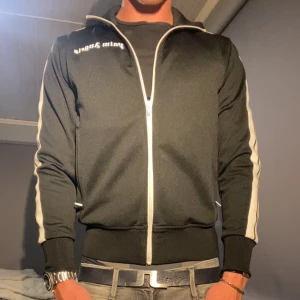 Palm Angels svart track jacket - Svart track jacket från Palm Angels med vita ränder längs ärmarna och dragkedja framtill. Jackan har ribbade muddar, ståkrage och logga på bröstet. Perfekt för en sportig och trendig look.