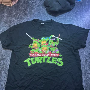Svart Teenage Mutant Ninja Turtles t-shirt - Cool svart t-shirt med färgglatt Teenage Mutant Ninja Turtles-tryck på bröstet. Klassisk passform med rund hals och korta ärmar. Perfekt för dig som älskar retro och nördiga motiv. Materialet känns mjukt och bekvämt, troligen bomull.