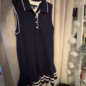 Marinblå ärmlös klänning Tommy Hilfiger - Snygg marinblå ärmlös klänning från Tommy Hilfiger med vit krage, knappar framtill och vita detaljer längs kanterna. Kjolen har sportiga ränder nertill och klänningen har en klassisk pikélook.