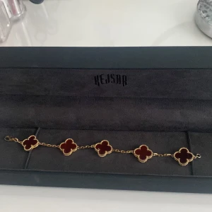 Guldigt armband med röda klöver från Kejsar - Snyggt armband från Kejsar med sex fyrklöverformade hängen i mörkröd färg, inramade av guldfärgade detaljer. Kedjan är tunn och elegant, perfekt för att ge din look en lyxig touch. Kommer i original ask med guldfärgad logga. Ordinarie pris 1000, pris går att diskuteras