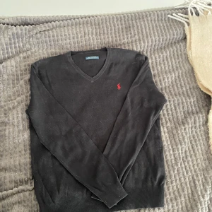 Svart v-ringad tröja från Polo Ralph Lauren - Svart v-ringad tröja från Polo Ralph Lauren med röd broderad logga på bröstet. Tröjan är långärmad och har ribbade muddar vid ärmslut och nederkant. Perfekt för dig som gillar klassisk och stilren look.