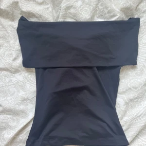 Svart axelbandslös tubtopp - Säljer en stilren svart tubtopp med bred kant upptill. Toppen är axelbandslös och har en tight passform som sitter snyggt på kroppen. Aldrig använd, prislappen är kvar.