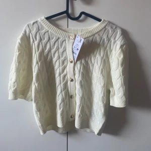 Vit kabelstickad kofta med kort ärm från gina - Supersöt vit kabelstickad kofta med korta ärmar och snygga guldfärgade knappar framtill. Koftan har rund halsringning och ribbade muddar. Perfekt för lager-på-lager och ger en clean, fräsch look. snabb affär får för 100