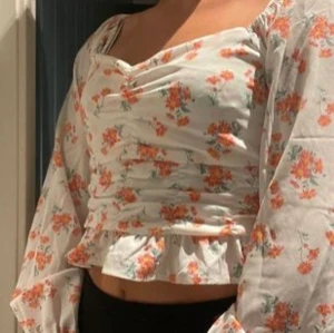 Blommig blus från Bik Bok med volang - Supersöt blommig blus från Bik Bok i vitt med orange och rosa blommor. Toppen har långärmade puffärmar, v-ringning, rynkade detaljer framtill och volangkant nertill. Perfekt för vår och sommar, och ger en romantisk vibe till din outfit.