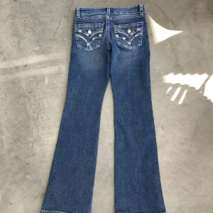 Blå bootcut jeans med broderade fickor - Snygga blå bootcut jeans med broderade bakfickor och paljett-detaljer. Jeansen har normal passform, klassisk femficksdesign och coola knappar på bakfickorna. Perfekta för dig som gillar lite extra detaljer på dina jeans. Gina young.