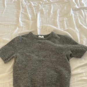 Grå stickad croppad tröja H&M - Grå stickad tröja från H&M med kort ärm och croppad passform. Enkel och stilren med rund halsringning och ribbade kanter. Perfekt att matcha med höga jeans eller kjol för en chill look. Aldrig använd. Pris kan diskuteras.