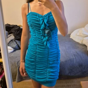 Turkos kortklänning med volangdetalj - Snygg turkos kortklänning med smala axelband och draperad volang framtill. Klänningen har rynkade sömmar längs hela framsidan och en dekorativ blomdetalj i tyget. Perfekt för dig som vill sticka ut med färg och coola detaljer. Storlek M men passar även S. 