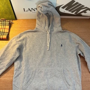 Grå hoodie från Polo Ralph Lauren - Klassisk grå hoodie från Polo Ralph Lauren med broderad blå logga på bröstet. Tröjan har huva med snörning, känguruficka och långa ärmar. Perfekt för en chill och stilren look. Mjuk bomullskänsla och relaxed fit.