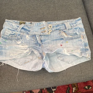 River Island ljusblå jeansshorts - Snygga ljusblå jeansshorts från River Island med slitna detaljer och råa kanter. Shortsen har flera knappar i midjan och små broderade färgprickar. Perfekta för sommaren och ger en cool, avslappnad vibe. Priset kan diskuteras💖