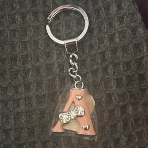 Nyckelring med bokstaven A och strass - Cool nyckelring med ett stort A i persikofärg, dekorerad med små hjärtan och glittrande strassdetaljer. Bokstaven sitter i en transparent plast och har silvriga metalldelar. Perfekt accessoar för att göra din väska eller nyckelknippa personlig.