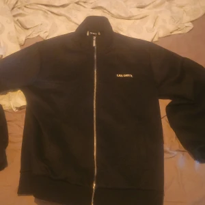 Svart fleecejacka från Les Deux - Svart fleecejacka från Les Deux med hel dragkedja framtill och diskret logga på bröstet. Jackan har hög krage och en clean, stilren look som passar perfekt till lager-på-lager. Enkel och snygg att slänga på sig när det är kyligt ute.