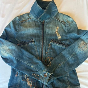 Y2K jeans jacka  - Snygg blå jeansjacka med coola nitar och stenar i brons och guld på både fram- och baksida. Jackan har dragkedja framtill, höga fickor med dragkedja och markerad krage. Unika slitningar och detaljer ger jackan en edgy vibe.