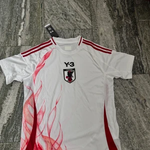 Japan Y-3 fotbollströja Adidas M - Japan landslagströja designad av Y-3 och Adidas. Vit bas med röda detaljer, tre röda ränder på axlarna och unikt rödflammigt mönster på fram- och baksidan. Broderad Japan-logga och Y-3-tryck på bröstet. Tillverkad i lätt, ventilerande material.
