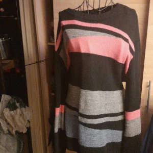 Randig stickad klänning från Xlnt - Mysig stickad klänning från Xlnt i svart, grått och rosa med breda ränder. Modellen är långärmad och har en rak, loose passform som är perfekt för kyligare dagar. Klänningen är knälång och har en enkel rund halsringning.