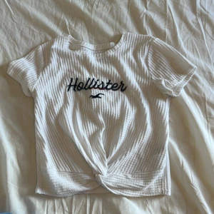 Vit ribbad topp från Hollister - Vit ribbad topp från Hollister med broderad logga i svart framtill. Kortärmad modell med rund hals och snygg knutdetalj längst ner. Perfekt för en chill och stilren look, passar till både jeans och kjol.