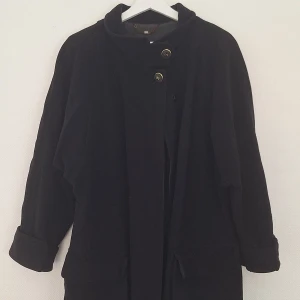 Svart kappa från Cirstein i kashmirull - Stilren svart kappa från Cirstein i lyxig mix av kashmir och ull. Kappan har hög krage, snygga guldknappar. Perfekt för dig som gillar klassisk och clean look med en twist.