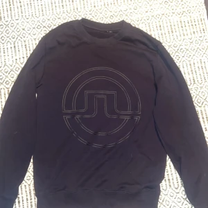 Svart sweatshirt från J.Lindeberg - Svart sweatshirt från J.Lindeberg med stort, stilrent logotryck framtill. Klassisk rund halsringning och långa ärmar. Perfekt för dig som gillar enkel men snygg design. Mjuk och skön bomullskvalitet. Skick 10/10 lite solbränd beroende på hur ljuset strålar på den men annars felfri 