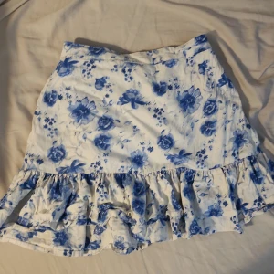 Blommig volangkjol från Lindex - Supersöt kort kjol från Lindex med blått blommönster på vit botten. Kjolen har volang nertill och resår i midjan för extra bekvämlighet. Perfekt för dig som gillar en romantisk och somrig stil. Väldigt stretchig. Passar mig som har 164 i storlek/xs-s 