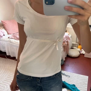 Vit t-shirt med rosett från 157 - Supersöt vit t-shirt från 157 i bomull med korta ärmar och en smal satinrosett under bysten. Enkel och clean design som passar perfekt till jeans eller kjol. T-shirten har en feminin passform och känns riktigt fräsch. Satin bandet har jag sytt på själv! 🥹❣️