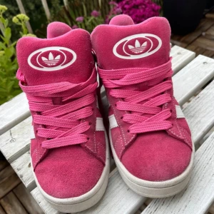 Adidas Campus rosa sneakers i mocka - Snygga Adidas Campus sneakers i en stark rosa färg med vita detaljer och klassiska tre ränder på sidan. Ovandel i mjuk mocka, vita loggor och platt sula. Perfekta för dig som vill sticka ut med färg och streetstyle.