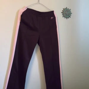 Svarta mjukisbyxor Hugo Boss med rosa revärer - Svarta mjukisbyxor från Hugo Boss med breda ljusrosa revärer längs sidorna. Byxorna har elastisk midja, dragkedjefickor och en relaxed passform. Perfekta för chill dagar eller när du vill ha en sportig vibe.