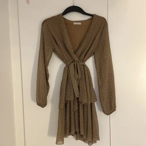 Beige omlottklänning med volang - Supersöt beige kortklänning i omlottmodell med knyt i midjan och volangdetaljer. Långärmad och lätt transparent tyg med små prickar. Tillverkad i Italien av mjuk polyester med lite stretch. Perfekt för dig som gillar en feminin och flowy stil.