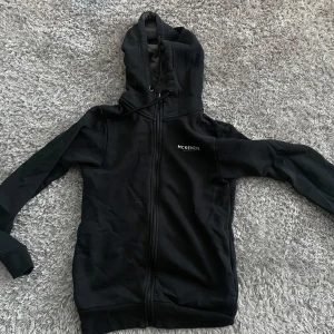 Svart hoodie från McKenzie med dragkedja - Svart hoodie från McKenzie med hel dragkedja och huva med snörning. Mjuk insida och diskret logga på bröstet. Perfekt för chill dagar eller när du vill ha en enkel och stilren look. Denna är använd några gånger och är i storlek XS