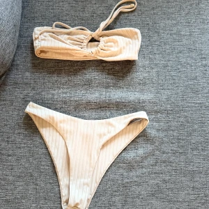 Beige randig bikini med kjol - Supersnygg bikini i ljusbeige med diskreta ränder. Bandeau-topp med knytning, samt matchande bikinitrosa med hög benskärning. Till setet hör även en kort kjol med knyt i sidan. Perfekt för stranden eller poolen!