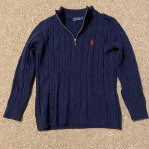 Marinblå stickad half zip från Polo Ralph Lauren - Säljer en marinblå stickad tröja från Polo Ralph Lauren med half zip och klassiskt kabelstickat mönster. Tröjan har lång ärm, ribbade muddar och röd broderad logga på bröstet. Perfekt för dig som gillar preppy och stilren look.
