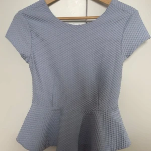 Ljusblå peplum topp med öppen rygg - Säljer en ljusblå peplum topp med korta ärmar och cool struktur i tyget. Toppen har en snygg öppen rygg med breda band och en markerad midja som ger en feminin siluett. 