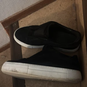 Svarta sneakers från Axel Arigato - Stilrena svarta sneakers från Axel Arigato med vit platt sula och diskret logga på sidan. Ovandelen är i mocka och skon har en minimalistisk slip-on design utan snörning. Perfekta för dig som gillar clean och modern streetstyle.