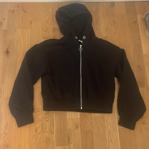 Svart hoodie med dragkedja från Lindex - Svart hoodie från Lindex med hel dragkedja framtill och huva. Tröjan har långa ärmar och breda ribbade muddar vid ärmslut och midja. Perfekt för en chill och avslappnad stil. Materialet känns mjukt och bekvämt, passar till många outfits.