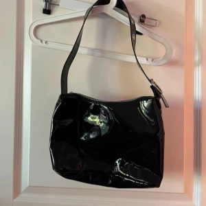 Svart glansig axelväska med dragkedja - Snygg svart axelväska i glansigt syntetmaterial med enkel design och dragkedja upptill. Perfekt storlek för det viktigaste och har en bred axelrem med metallspänne. Väskan har en modern vibe och passar till många olika outfits.