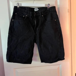 Svarta jeansshorts från 157 - Svarta jeansshorts från 157 med klassisk femficksdesign och knappgylf. Modellen är rak och lite längre i benen, perfekt för en chill och stilren look. Passar dig som gillar enkel och tidlös stil.