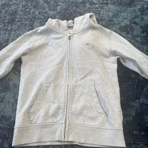 Grå hoodie från Gant med dragkedja - Säljer en ljusgrå hoodie från Gant med dragkedja framtill och två stora fickor. Tröjan har huva, ribbade muddar och en liten broderad Gant-logga på bröstet. Perfekt för lager-på-lager och chill dagar.