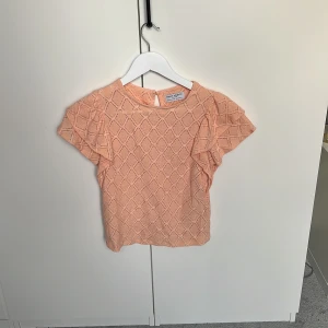 Persikofärgad blus från Holly & Whyte - Supersöt persikofärgad blus från Holly & Whyte med korta volangärmar och ett snyggt rutmönster i strukturen. Blusen har rund halsringning och knapp i nacken. Perfekt för dig som gillar en feminin och fräsch stil.