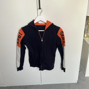 Svart och orange hoodiejacka från Gant - Svart hoodiejacka från Gant med orange huva och detaljer, samt vita partier på ärmarna. Snygg dragkedja framtill och tryck med GANT och 1949 på ärmarna samt 'NEW HAVEN' på ryggen. Perfekt för en sportig och chill look.