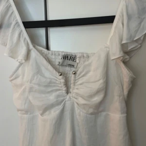 Vit blus med volangärm från Aware - Supersöt vit blus från Aware med volangärmar och snygg v-ringning med rynk och små silverfärgade detaljer framtill. 