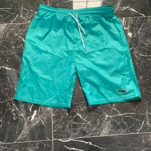 Turkosa badshorts från Lacoste - Snygga turkosa badshorts från Lacoste med klassisk krokodillogga på benet. Shortsen har elastisk midja med vit snörning och är tillverkade i ett lätt syntetmaterial som torkar snabbt. Perfekta för stranden eller poolen i sommar.