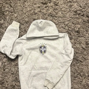 Ljusgrå hoodie med Saint-logga Brazil tröja  - Ljusgrå hoodie från Saint med broderad sköldlogga på bröstet, fem gröna stjärnor ovanför och klassisk känguruficka. Tröjan har huva och långa ärmar, perfekt för en chill och sportig stil.